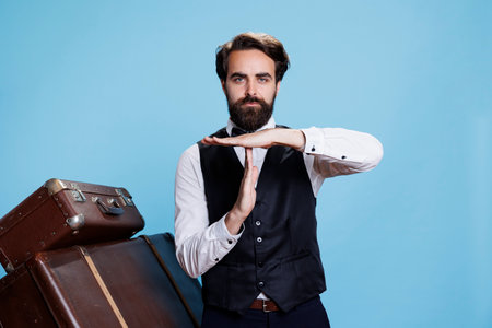 Hotel porter shows timeout symbol on camera, asking for a break working in hospitality and travel industry. Classy doorman in suit and tie indicating pause sign with hands, professional job.の写真素材