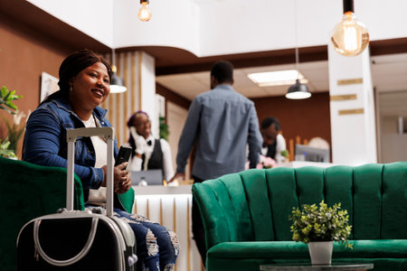Happy young African American woman sitting at hotel lobby with suitcase holding smartphone and smiling, using phone for mobile self check-in. Black female tourist with luggage waiting for roomの写真素材