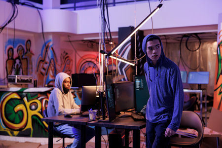 Suspicious hacker looking away after breaking into server and doing illegal activity. Young asian criminals in hood leaving abandoned warehouse after breaching database and stealing dataの写真素材