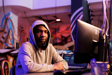 African american hacker doing illegal activities on computer and working late portrait. Criminal in hood breaking website with ddos attack and looking at camera in abandoned warehouseの写真素材