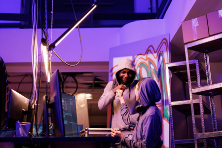 Cheerful hackers bumping fists while hacking online server successfully. Internet criminals greeting with friendly gesture while breaking law and coding malicious software on computerの写真素材