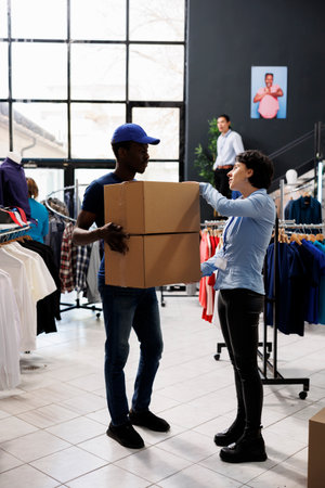 Store worker explaining customer shipping detalis to african american courier in modern boutique. Man wearing delivery uniform, holding cardboard boxes with online orders in shopping centreの写真素材
