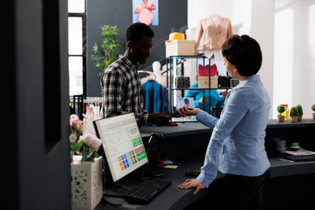 Stylish customer paying for fashionable clothes with credit card using payment terminal during fashion shopping session in modern boutique. African american man buying formal wearの写真素材