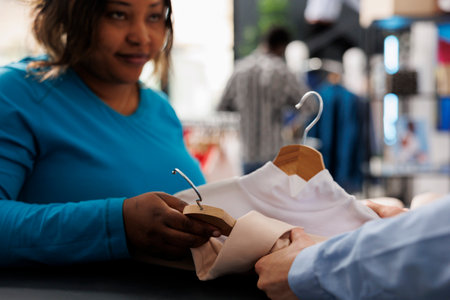 African american woman shopping for casual wear, holding multiple shirts in modern boutique. Stylish customer standing at counter desk, discussing items fabric with store employee. Fashion conceptの写真素材