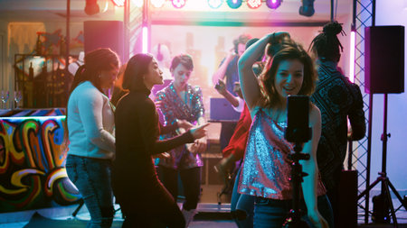 Happy girl recording vlog for internet, using mobile phone at discotheque on dance floor. Female influencer filming cool video of people dancing together and jumping, disco event. Handheld shot.の写真素材