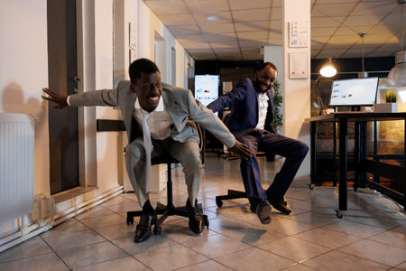 Playful businessmen riding desk chair during sports race competition during break time in modern startup office. Cheerful african american employees having fun, enjoying doing moving activityの写真素材