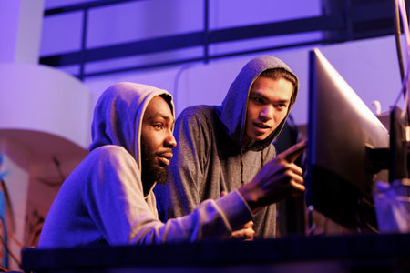 Hackers exchanging ideas and pointing at screen while planning phishing fraud. African american and asian criminals in hoods checking confidential data after breaking into serverの写真素材