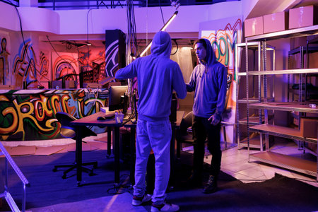 Two hackers discussing phishing attack in abandoned warehouse with graffiti. Young internet...