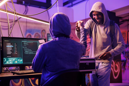Cheerful african american hacker giving fist bump to crime partner while working together in abandoned warehouse. Smiling men in hoods sharing friendly gesture after successful database breakingの写真素材