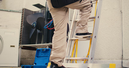 Ground level shot of capable electrician holding hand drill climbing folding ladder to do maintenance on rooftop hvac system. Seasoned wireman commissioned to checkup on condenserの写真素材