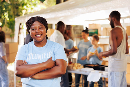 Enthusiastic black woman wearing blue t-shirt with arms crossed looks at camera. Portrait shot of african american female volunteer, ready to provide humanitarian aid to poor, needy and less fortunate.の写真素材