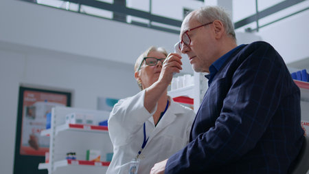 Elderly pharmacist in drugstore using electronic thermometer on senior man, checking temperature. Old patient and healthcare specialist during yearly examination, monitoring flu symptoms such as feverの写真素材