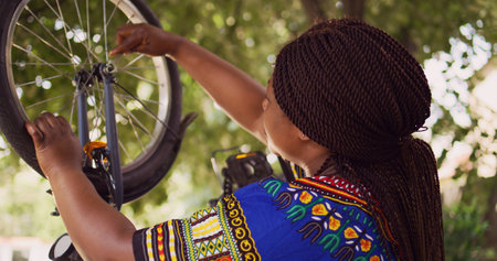 Healthy black woman mending her own bicycle using specialized equipment from toolkit in yard. Dolly zoom-in shot. Female african american cyclist repairing front bike wheel outdoors.の写真素材