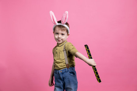 Positive cheerful preschooler playing in studio with a toy, feeling joyful and excited while he wears bunny ears. Happy young child posing against pink background, innocent little boy.の写真素材