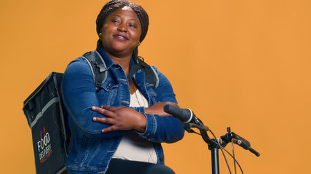 African american delivery woman with arms crossed resting on her bicycle. Young professional courier with backpack ready for environmentally friendly delivery service in urban neighborhood.の写真素材