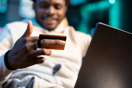 Close up shot of debit card in man hand adding payment method on website. Focus on credit card held by person in blurry background doing online shopping from neon lit apartmentの写真素材