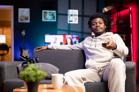 Man zapping through TV channels using remote, enjoying leisure time in living room used as home...