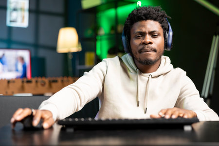 BIPOC man wearing headphones playing online multiplayer videogame on gaming PC, contending against...