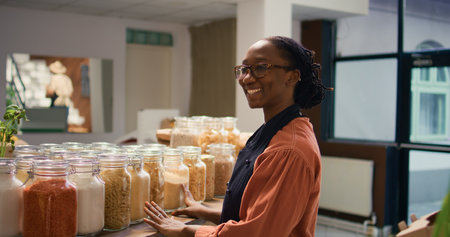 African american seller in supermarket refilling shelves with pasta or sauces, freshly harvested produce in crates. Small business owner in local neighborhood zero waste eco store.の写真素材