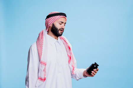 Muslim man dressed in cultural clothes holding medication and reading instruction on bottle. Person wearing traditional arabian robe and headscarf checking vitamins packageの写真素材