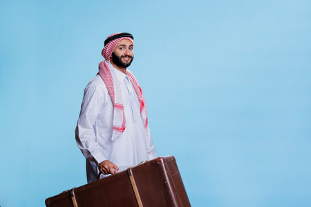 Smiling muslim traveler holding vintage leather suitcase and looking at camera with cheerful expression. Happy arab dressed in traditional islamic clothes carrying baggage studio portraitの写真素材