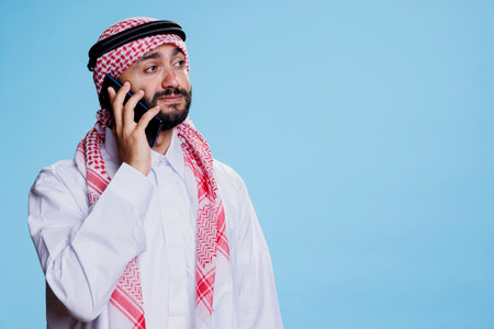 Muslim man dressed in islamic clothes, answering call and having conversation on mobile phone. Arab person dressed in traditional thobe and ghutra while talking on smartphoneの写真素材