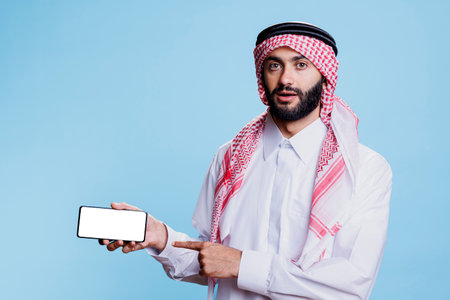 Muslim man holding smartphone with white screen in horizontal mode and pointing with finger. Arab person in traditional clothes showing empty touchscreen and looking at cameraの写真素材