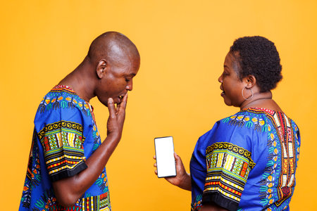 Woman showing smartphone with empty white screen with copy space to shocked man. Surprised person covering mouth with hand while looking at mobile phone with blank touchscreen mockup for appの写真素材