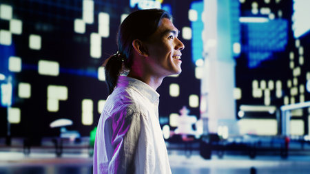 Asian man wandering around city streets at night, looking upwards to surrounding condominiums in empty urban center with no traffic. Cheerful businessman taking a walk to clear his mindの写真素材