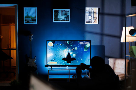 Player participates in online roleplaying firing match, hooking console through TV and enjoying nighttime gaming activity with friends. Male student is playing shooter video games.の写真素材