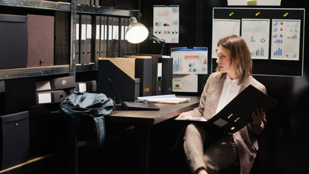 Determined private detective grabs files from shelves to conduct thorough investigation. Caucasian female agent with laptop and research papers, analyzes evidence and gathers information. Tripod shot.の写真素材