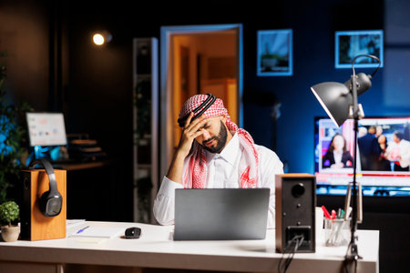 Frustrated businessman in Arabic clothing sitting at an office desk uses a laptop for online research. Young Muslim guy with a minicomputer on the table appears exhausted with a headache.の写真素材