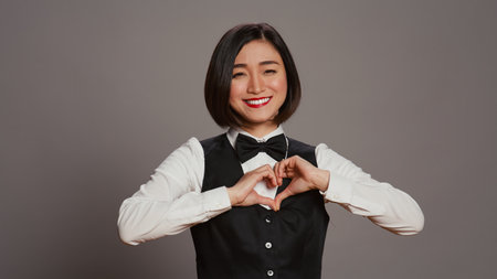 Hotel administrator shows heart shape symbol on camera, expressing romantic gesture and being flirty. Asian receptionist presenting true honest feelings, does romance sign in studio. Camera B.の写真素材