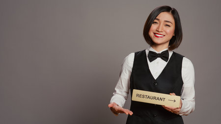 Asian hotel concierge holding restaurant sign to indicate direction, pointing towards dining area. Receptionist assisting clients to enjoy all amenities, stands over grey background. Camera B.の写真素材