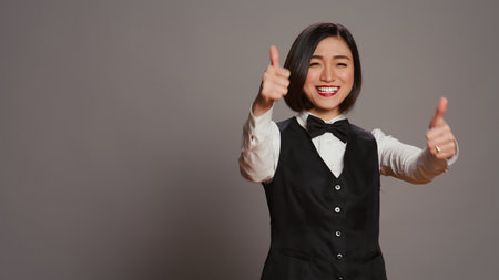 Asian reception staff doing thumbs up symbol on camera, showing approval and positive sign over grey background. Hotel receptionist giving like and okay gesture, showing agreement. Camera B.の写真素材