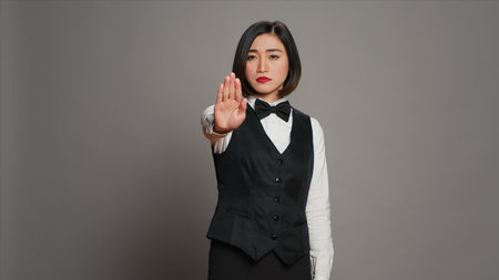 Asian administrator showing stop sign with palm raised in studio, presenting negative symbol and expressing disapproval. Woman receptionist doing no gesture, being displeased. Camera A.の写真素材