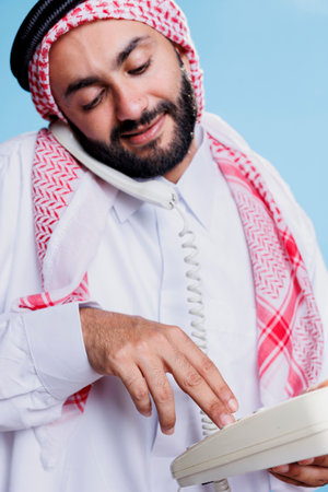 Muslim man wearing traditional headscarf dialing number on landline phone. Arab person dressed in...