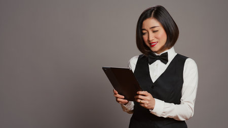 Asian hotel administrator browsing online website to check all reservations or room bookings, smiling and being confident. Receptionist using tablet in studio, hospitality industry. Camera B.の写真素材