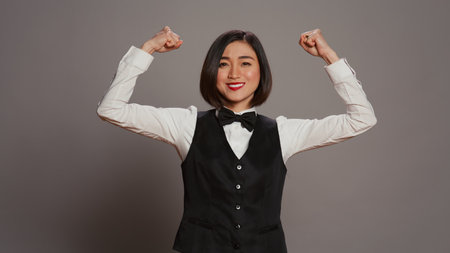 Asian hotel concierge flexing arms muscles in grey studio, showing off her strength and power in front of camera. Receptionist with bow and uniform standing with biceps flexed. Camera B.の写真素材