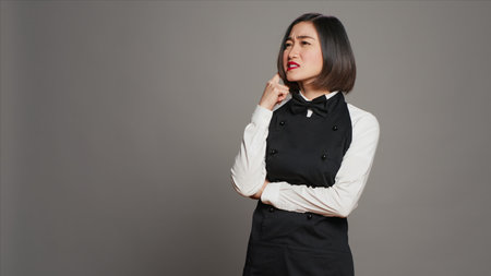 Restaurant hostess being thoughtful over grey background, thinking about new ideas to develop the menu for clients. Asian woman waitress feeling pensive, imagination in studio. Camera A.の写真素材