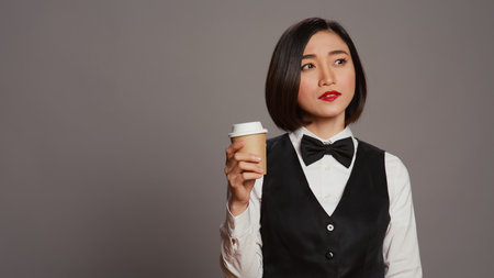 Asian receptionist serving coffee cup in front of camera, taking a break and enjoying hot refreshment. Hotel administrator drinking caffeine beverage, tourism concept. Camera B.の写真素材
