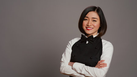 Restaurant hostess posing with confidence in studio, standing with arms crossed over grey background. Asian waitress barista with gourmet serving expertise smiling on camera. Camera B.の写真素材
