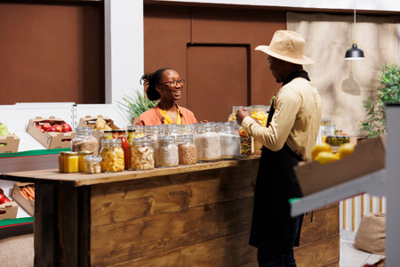 Male shop owner discussing fresh fruits and vegetables at local market with a smiling black woman. Seller offers organic pantry staples and plastic-free bulk products, promoting healthy produce.の写真素材