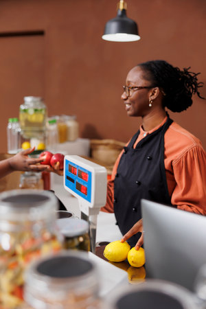 Black woman assists a customer in a grocery store, promoting locally grown and organic products. They communicate and use technology for an efficient checkout process in the eco-friendly market.の写真素材