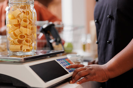 Up-close image capturing person using measuring scale, checking weight of container filled with organic bio goods. Black individual weighing glass jar of pasta at the checkout counter.の写真素材