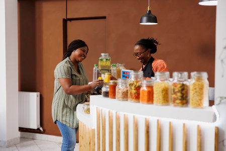 African American customer discusses eco friendly shopping with a store clerk at the checkout while handing over jars of cereal and honey for scanning. Black woman purchasing fresh bio food products.の写真素材