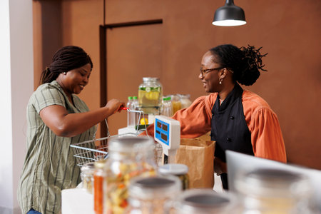 African American female customer unloading her shopping basket at the cashier counter. Friendly black woman vendor with a smile, assisting shopper with her farm grown food groceries.の写真素材