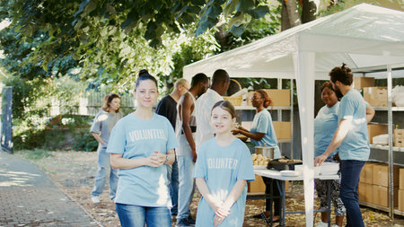 Portrait of a woman and daughter taking part in food bank program to fight hunger. Blue-shirted Caucasian volunteers are ready to help less fortunate while glancing into camera. Handheld shot.の写真素材