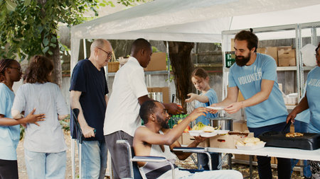 Humanitarian aid team providing assistance to the handicapped, poor and homeless people. Charity workers giving away free food to black man in wheelchair meeting his needs while others wait in line.の写真素材