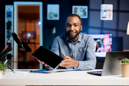 Influencer in home studio using recording tech to capture live broadcast, reviewing tablet. BIPOC online show host filming electronics haul, presenting digital device to viewersの写真素材